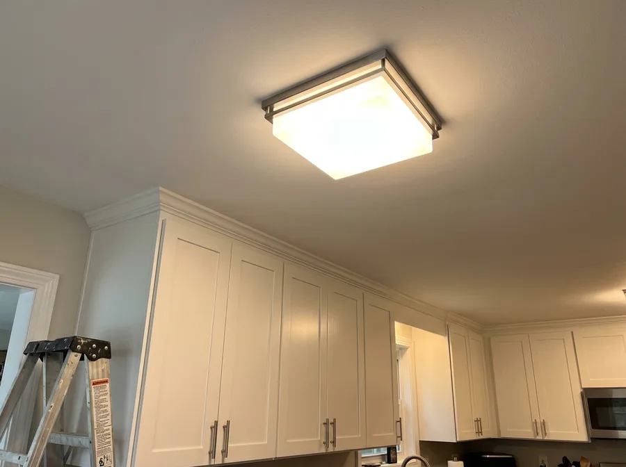A newly installed ceiling light fixture mounted flush and switched on, seen on a slight up-angle against a bright white ceiling above a home kitchen cabinet run, the electrician's folded step ladder just visible at the frame edge